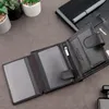 An open black leather wallet is displayed on a dark gray surface, with some small potted plants and a notebook in the background. Produktbild-Vorschau