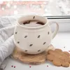 A mug of hot chocolate sits on a wooden coaster next to a knitted blanket, with snowflakes falling outside a window. Produktbild-Vorschau