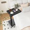 A C-shaped side table with a marble-like black top is placed next to a bed. The table holds a vase of flowers, a bowl of cherries, a cup of coffee, and a smartphone. The scene is set in a bedroom with white bedding and pillows. Produktbild-Vorschau