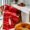 A cozy scene with a red and white patterned electric heated blanket draped over a wicker chair, next to a shelf with a mug and a small Christmas tree. Podgląd zdjęcia produktu