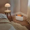 A white humidifier with a flame effect is placed on a wooden floor next to a bed and a stool with a lamp. Produktbild-Vorschau
