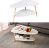 A white two-tiered oval coffee table with wooden legs is shown against a white background. The second image shows the same table in a living room setting with books, fruit, and a plant on it. Voir les images du produit