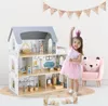 A young girl in a pink dress and crown stands next to a large wooden dollhouse, with a pink bear chair to her right. Wooden blocks are scattered on the floor in front of the dollhouse. A rose gold glittery banner hangs on the wall above. Produktbild-Vorschau