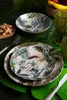 A table setting with tropical-themed plates, glasses, and a bowl of peanuts, surrounded by large green leaves. Produktbild-Vorschau