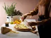 A person is using a wooden pizza cutter on a pizza, with ingredients and herbs in the background. Produktbild-Vorschau