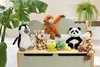 A collection of stuffed animals, including a panda, giraffe, monkey, leopard, penguin, and turtle, are arranged on a light green toy chest. A large green plant is visible in the background. Produktbild-Vorschau