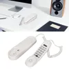 A white corded telephone with its base and handset shown separately on a white surface, with a computer and speaker in the background. Náhled obrázku produktu
