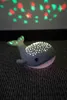 A whale-shaped night light projects stars onto a wall and sits on a textured surface. Produktbild-Vorschau