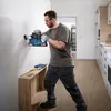 A man is using a cordless hammer drill to drill into a white wall in a room with wooden floors and kitchen cabinets in the background. Produktbild-Vorschau