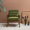 A green velvet armchair with a wooden frame is placed in a room with a brown wall and a patterned rug. Produktbild-Vorschau