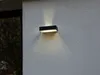 A black outdoor wall light fixture is mounted on a textured gray wall. The light emits a warm glow upwards and downwards. Produktbild-Vorschau