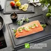A raw salmon fillet is prepared on a black cutting board, garnished with lemon slices and herbs, with other ingredients like tomatoes and lemons in the background. Podgląd zdjęcia produktu