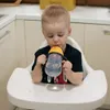 A young child sits in a high chair and drinks from a sippy cup. Anteprima dell’immagine prodotto