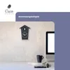 A cuckoo clock is displayed on a wall above a desk with a computer monitor, books, and a mug. Produktbild-Vorschau