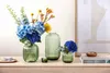 Three green glass vases with floral arrangements on a wooden surface, next to books and a small bowl. Náhled obrázku produktu