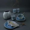A table setting with blue ceramic dishes, cutlery, and a vase, arranged with dried plant stems. Náhľad obrázku produktu