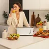 A woman drinks orange juice in a kitchen with a toaster and breakfast items on the counter. Voir les images du produit