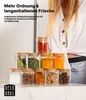 Klare Glasgewürzgläser mit Bambusdeckeln, pyramidenförmig gestapelt, gefüllt mit verschiedenen Gewürzen, neben einer Person in einem weißen Hemd Voir les images du produit