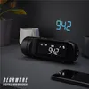 A digital alarm clock with a projector is placed on a dark surface next to a smartphone, with plants and decorative pots in the background. Náhľad obrázku produktu