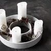 A circular candle holder with four white pillar candles, pine cones, and dark decorative balls on a dark wooden floor. Voir les images du produit