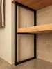 A close-up view of a black metal shelf frame with wooden shelves, set against a concrete and tiled wall. Produktbild-Vorschau