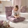 A young girl is sitting on a purple floor chair with a rainbow pattern, holding a small book. A wooden toy chicken is on the floor in front of her. A laundry basket with a pink towel is in the background. Anteprima dell’immagine prodotto