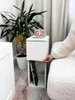 A person's hand is shown reaching for the drawer of a white bedside table next to a bed. A plant and a decorative mug are also visible. Náhľad obrázku produktu