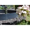 A close-up view of a black plastic garden edging around a wooden post, with gravel and blurred flowers in the foreground and background. Produktbild-Vorschau