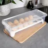 A clear plastic egg container with 12 eggs inside, placed on a wooden cutting board on a kitchen counter. Podgląd zdjęcia produktu