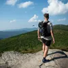 A person wearing a backpack stands on a rocky outcrop overlooking a forested mountain range. Anteprima dell’immagine prodotto