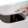 A close-up shot of a person's finger pressing down on the lid of a gray and white dual compartment trash can. The lid is slightly ajar, revealing the dark interior of one compartment. A mint green section is visible in the background. Produktbild-Vorschau