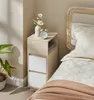 A narrow bedside table with two white drawers and an open shelf sits next to a bed with a woven headboard. Produktbild-Vorschau