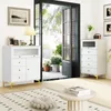 A modern living room with two white storage cabinets, one on the left and one on the right, with gold legs. A large glass door opens to a patio with plants. A rug and a round coffee table are in the foreground. Produktbild-Vorschau