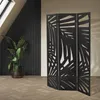 A black, three-panel room divider with a leaf-like cut-out pattern is shown in a room with a large potted plant and a slatted wall. Náhled obrázku produktu