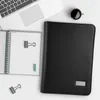 A black A4 conference folder, a notebook, a laptop, and a cup of coffee are arranged on a white surface. Produktbild-Vorschau