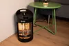 A black infrared heater is placed on a wooden floor next to a green side table and a sofa. Produktbild-Vorschau