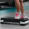 A person's legs and feet are shown stepping onto a black aerobic stepper in a gym. Podgląd zdjęcia produktu
