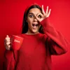 A woman wearing a red sweater and headband holds a red mug with 'Zilan' written on it. She is looking at the camera with one eye covered by her fingers in a playful gesture. The background is solid red. Podgląd zdjęcia produktu