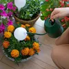 A person is watering plants using a green watering can and a white watering globe in a pot of orange marigolds. Other plants and flowers are visible in the background. Anteprima dell’immagine prodotto