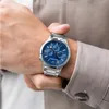 A man's wrist wearing a silver watch with a blue dial, partially covered by a light brown jacket sleeve. Náhľad obrázku produktu
