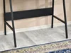 Close-up view of the black metal legs and support bar of a desk, with a patterned rug and light-colored floor visible. Náhled obrázku produktu