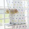 A close-up view of sheer curtains with colorful pom-pom accents, hanging in front of a window with a vase of flowers on a shelf. Produktbild-Vorschau