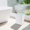 A white dual-compartment trash can sits next to a white bathtub in a well-lit bathroom with a large window overlooking greenery. Produktbild-Vorschau