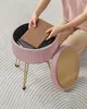 A person is placing a book into a pink velvet storage ottoman with gold legs. Náhled obrázku produktu