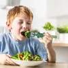 A young boy is eating broccoli from a bowl with a dinosaur-shaped fork. Produktbild-Vorschau