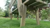 A wooden garden bench is shown in a grassy backyard with plants and a fence in the background. Produktbild-Vorschau