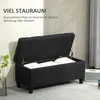 A black storage bench with its lid open, revealing white pillows inside. The bench is placed in a room with a plant and a speaker in the background. Text overlays describe the storage capacity. Náhled obrázku produktu