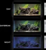 Three aquarium tanks are shown under different lighting conditions: Day, Daybreak, and Night. Each tank contains rocks and aquatic plants. A lighting fixture is shown to the right of each tank. Anteprima dell’immagine prodotto