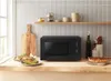 A black microwave oven is placed on a wooden countertop in a kitchen setting, with food items and kitchenware around it. Produktbild-Vorschau