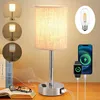 A modern LED table lamp with a linen shade and USB charging ports is displayed on a wooden desk next to an open book and glasses. The lamp is shown with multiple lighting options and charging capabilities for a phone and smartwatch. Produktbild-Vorschau